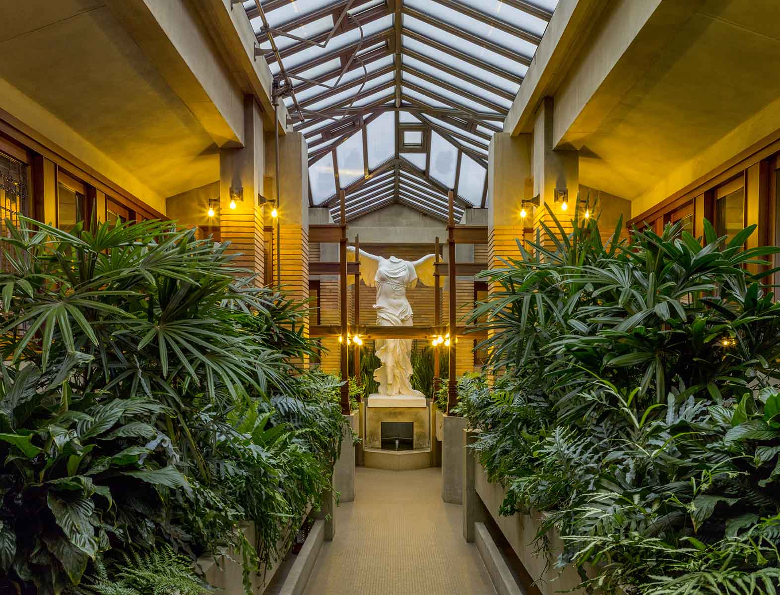 photo of Victory of Samothrace sculpture at the end of a path bordered by green plants in a glass-roofed conservatory with tan colors and warm lighting