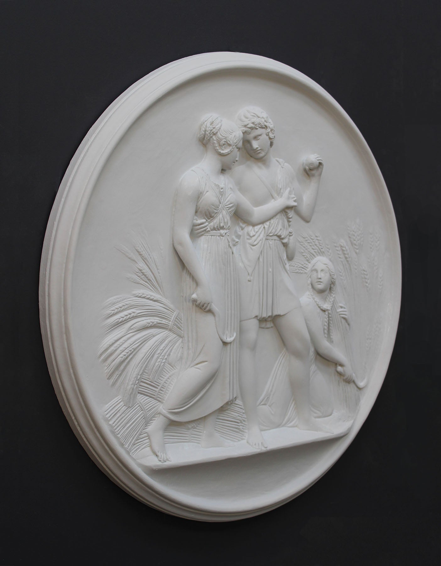 photo of white plaster cast relief sculpture of male and female figures walking through field of wheat and a female figure kneeling in the field cutting wheat against dark gray background