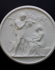 photo of white plaster cast relief sculpture of female figure with baby on lap, a standing male figure, and a dog against dark gray background