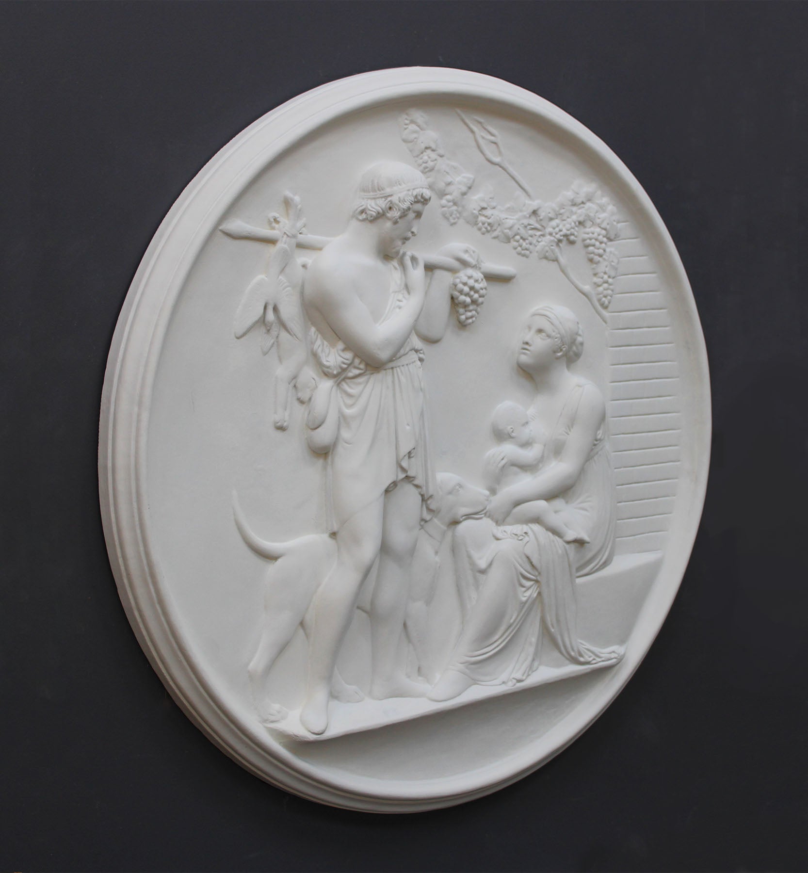 photo of white plaster cast relief sculpture of female figure with baby on lap, a standing male figure, and a dog against dark gray background