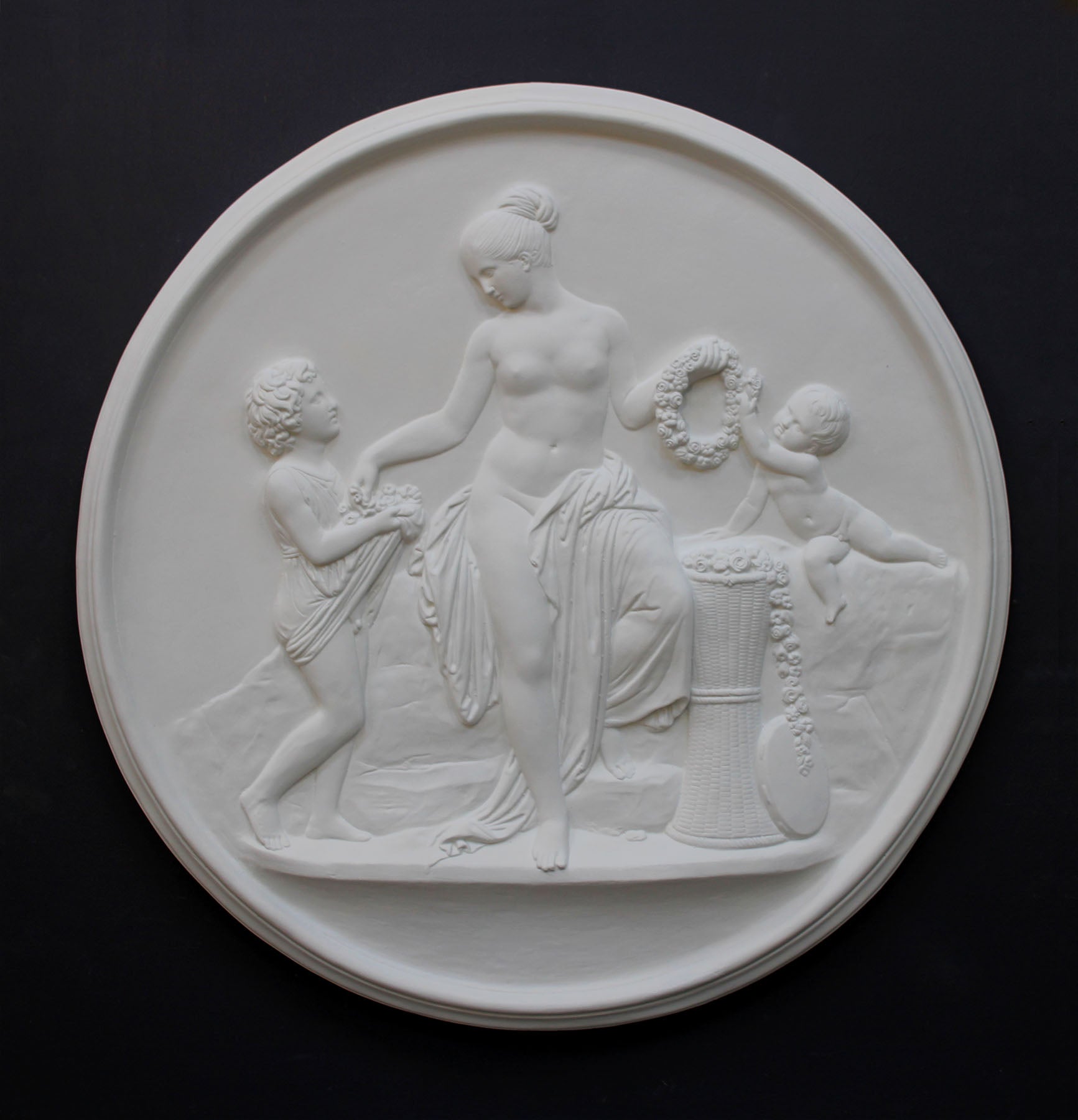 photo of white plaster cast relief sculpture of mostly nude female figure with two children and flowers and wreaths against dark gray background