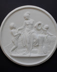 photo of white plaster cast relief sculpture of mostly nude female figure with two children and flowers and wreaths against dark gray background