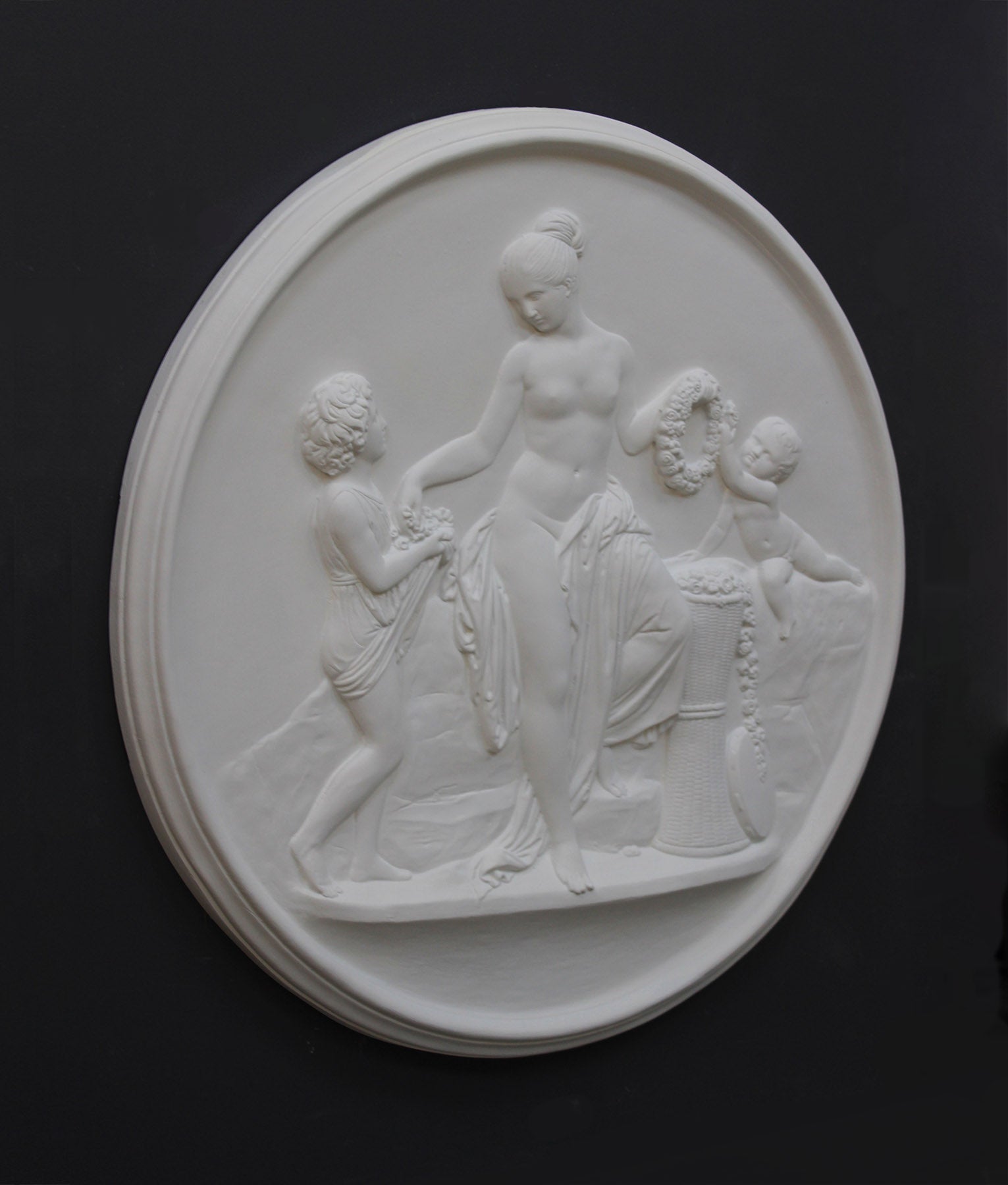 photo of white plaster cast relief sculpture of mostly nude female figure with two children and flowers and wreaths against dark gray background