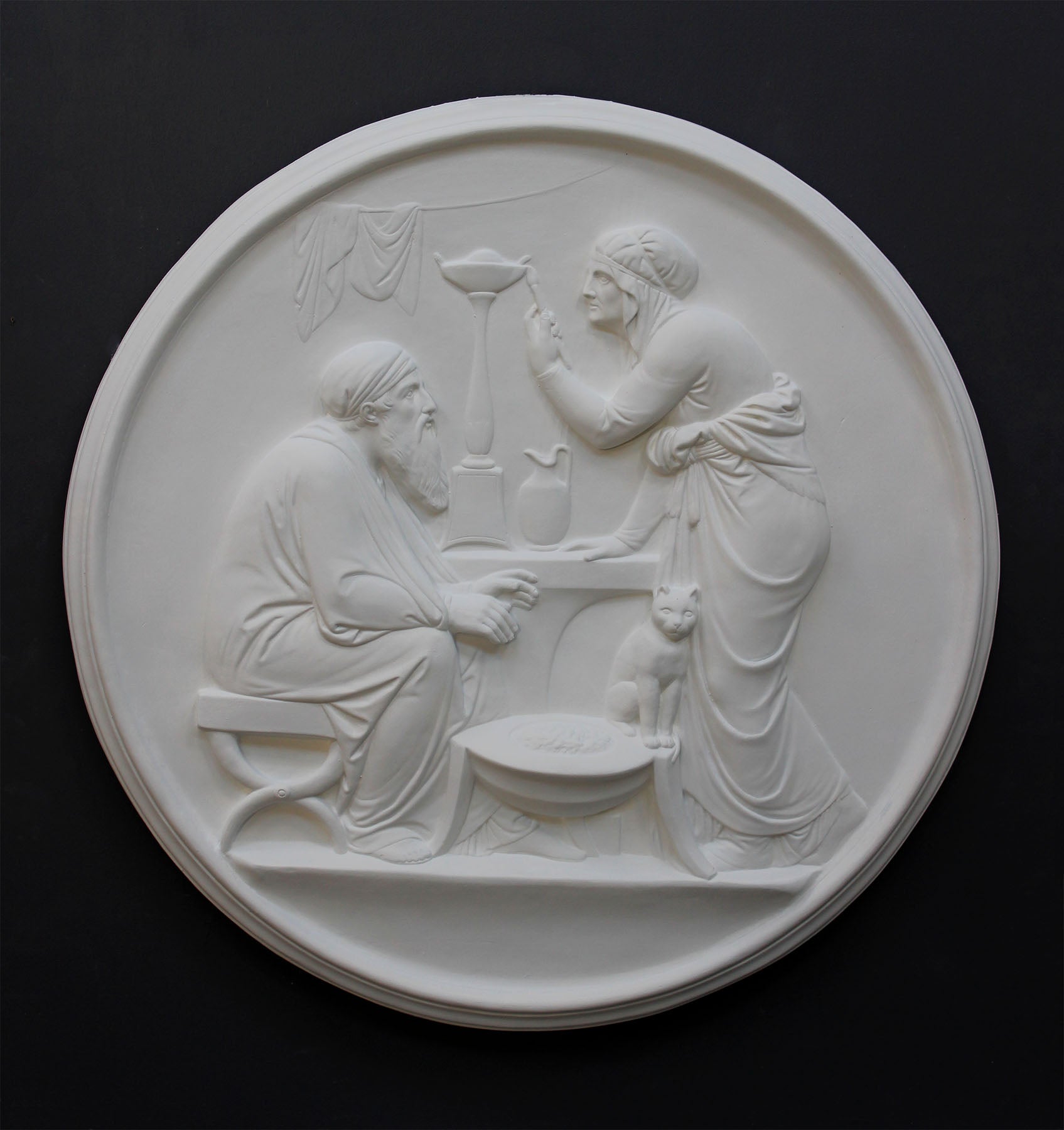 photo of white plaster cast relief sculpture of elderly male and female figures at a table and a cat against dark gray background