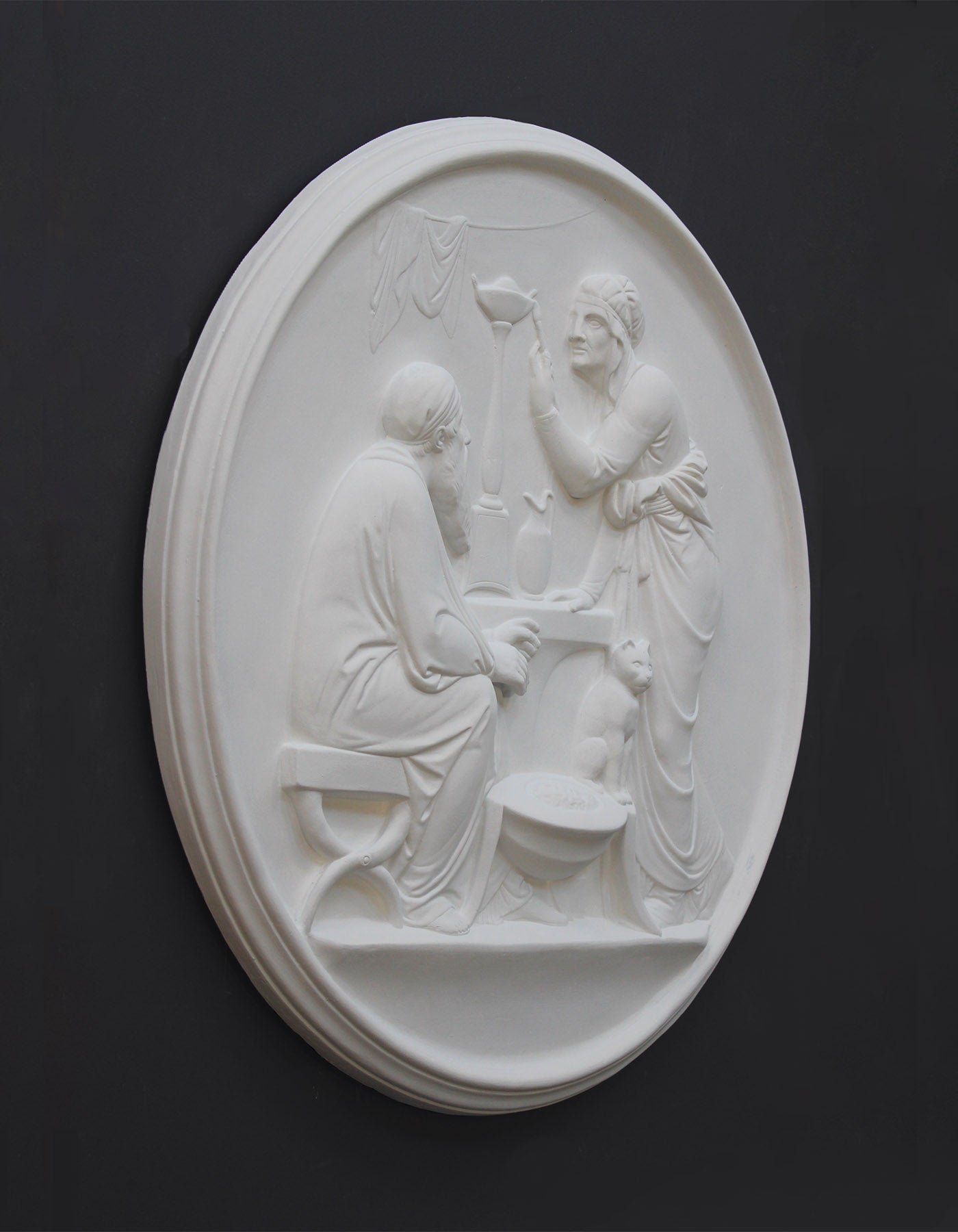 photo of white plaster cast relief sculpture of elderly male and female figures at a table and a cat against dark gray background