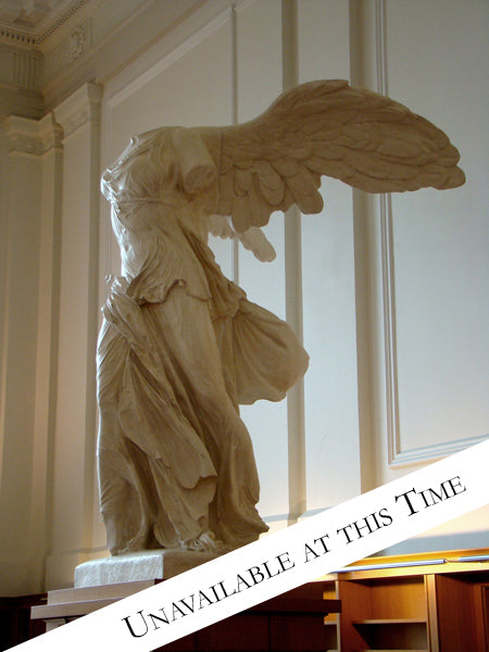 photo of plaster cast sculpture of winged, headless female figure with flowing drapery on top of a ribbed, square, wooden pedestal with white paneled walls behind