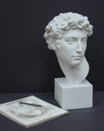 photo of white plaster cast bust sculpture of male with curly hair atop cube base with sketchbook and pencil beside it against gray background