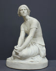 photo of white plaster cast sculpture of female, namely Joan of Arc, kneeling with hands on lap atop round pedestal on gray background