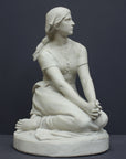 photo of white plaster cast sculpture of female, namely Joan of Arc, kneeling with hands on lap atop round pedestal on gray background