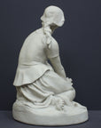 photo of back view of white plaster cast sculpture of female, namely Joan of Arc, kneeling with hands on lap atop round pedestal on gray background