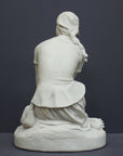 photo of back view of white plaster cast sculpture of female, namely Joan of Arc, kneeling with hands on lap atop round pedestal on gray background