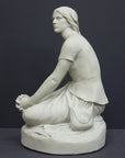 photo of white plaster cast sculpture of female, namely Joan of Arc, kneeling with hands on lap atop round pedestal on gray background