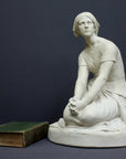 photo of white plaster cast sculpture of female, namely Joan of Arc, kneeling with hands on lap atop round pedestal with green book laid beside it on gray background