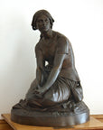 photo of bronze-colored plaster cast sculpture of female, namely Joan of Arc, kneeling with hands on lap on a wood pedestal with white wall behind