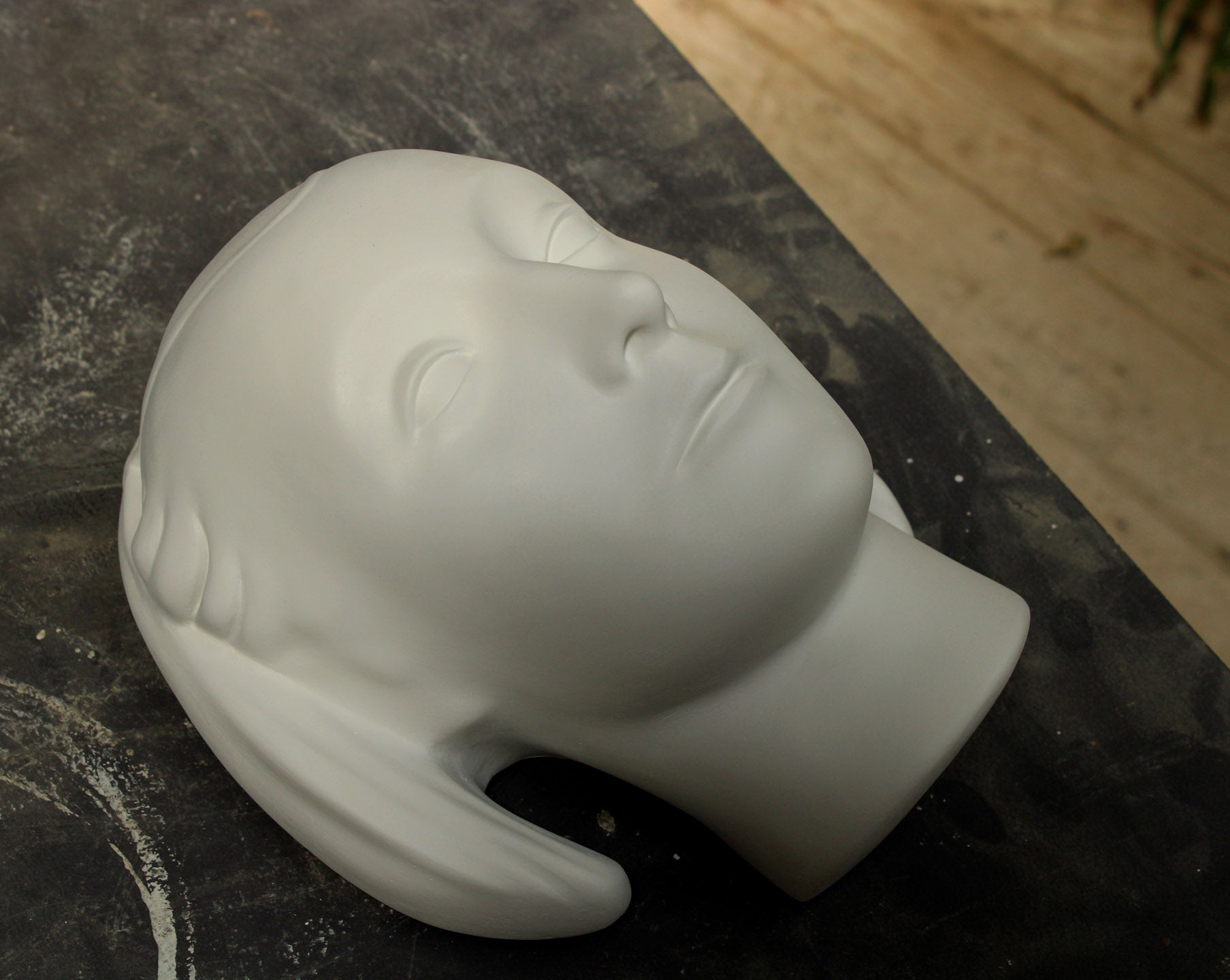 photo of white plaster cast sculpture mask of female with part-back hairdo set down on gray background with wood flooring visible