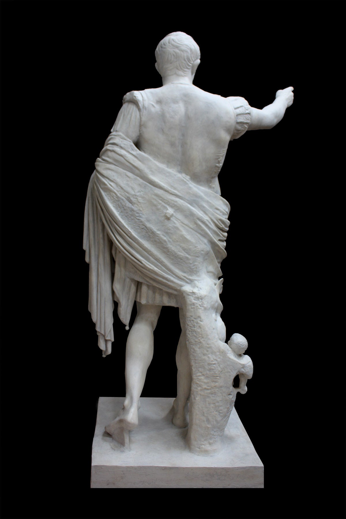 photo of reverse of white plaster cast sculpture of a man with armor and robes standing in contrapposto with his right arm raised, namely Augustus Caesar, against a black background