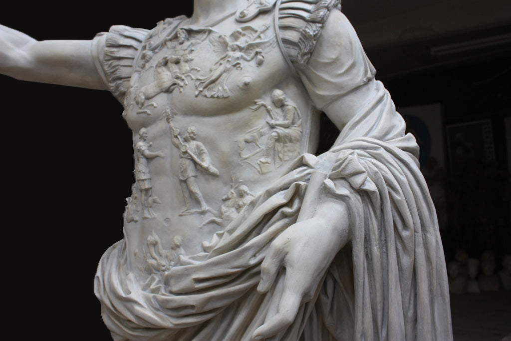 detail photo of armor from white plaster cast sculpture of a man with armor and robes standing in contrapposto with his right arm raised, namely Augustus Caesar, against a darkened background