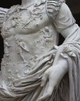 detail photo of armor from white plaster cast sculpture of a man with armor and robes standing in contrapposto with his right arm raised, namely Augustus Caesar, against a darkened background