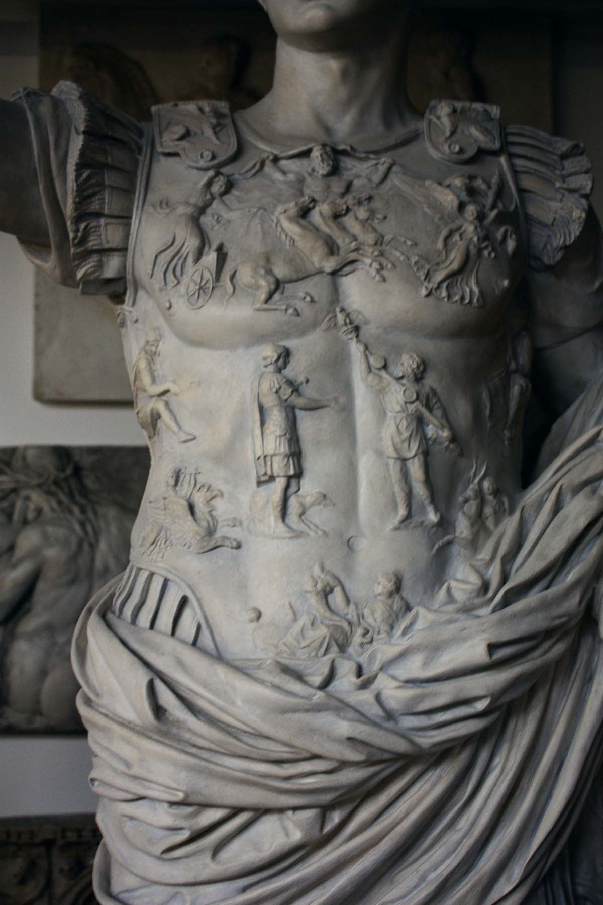 detail photo of armor from white plaster cast sculpture of a man with armor and robes standing in contrapposto with his right arm raised, namely Augustus Caesar, with relief sculptures hanging in gallery behind