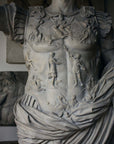 detail photo of armor from white plaster cast sculpture of a man with armor and robes standing in contrapposto with his right arm raised, namely Augustus Caesar, with relief sculptures hanging in gallery behind