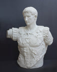 photo of white plaster cast sculpture of a man's torso with armor and robes with his right arm raised, namely Augustus Caesar, against a gray background