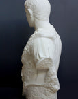 side view photo of white plaster cast sculpture of a man's torso with armor and robes with his right arm raised, namely Augustus Caesar, against a gray background