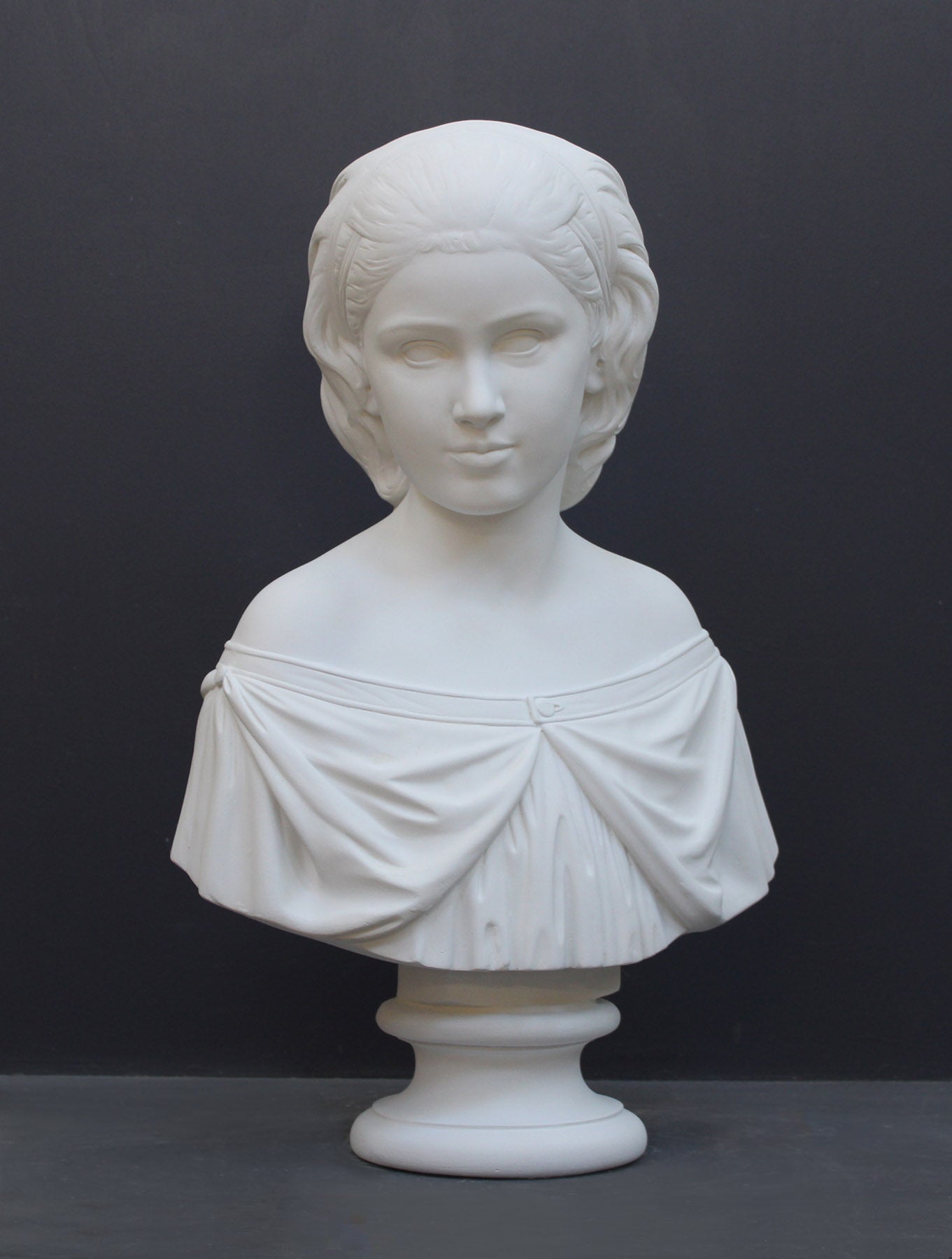 photo of white plaster cast bust sculpture of young woman with headband piece and in dress that falls off shoulders against a gray background