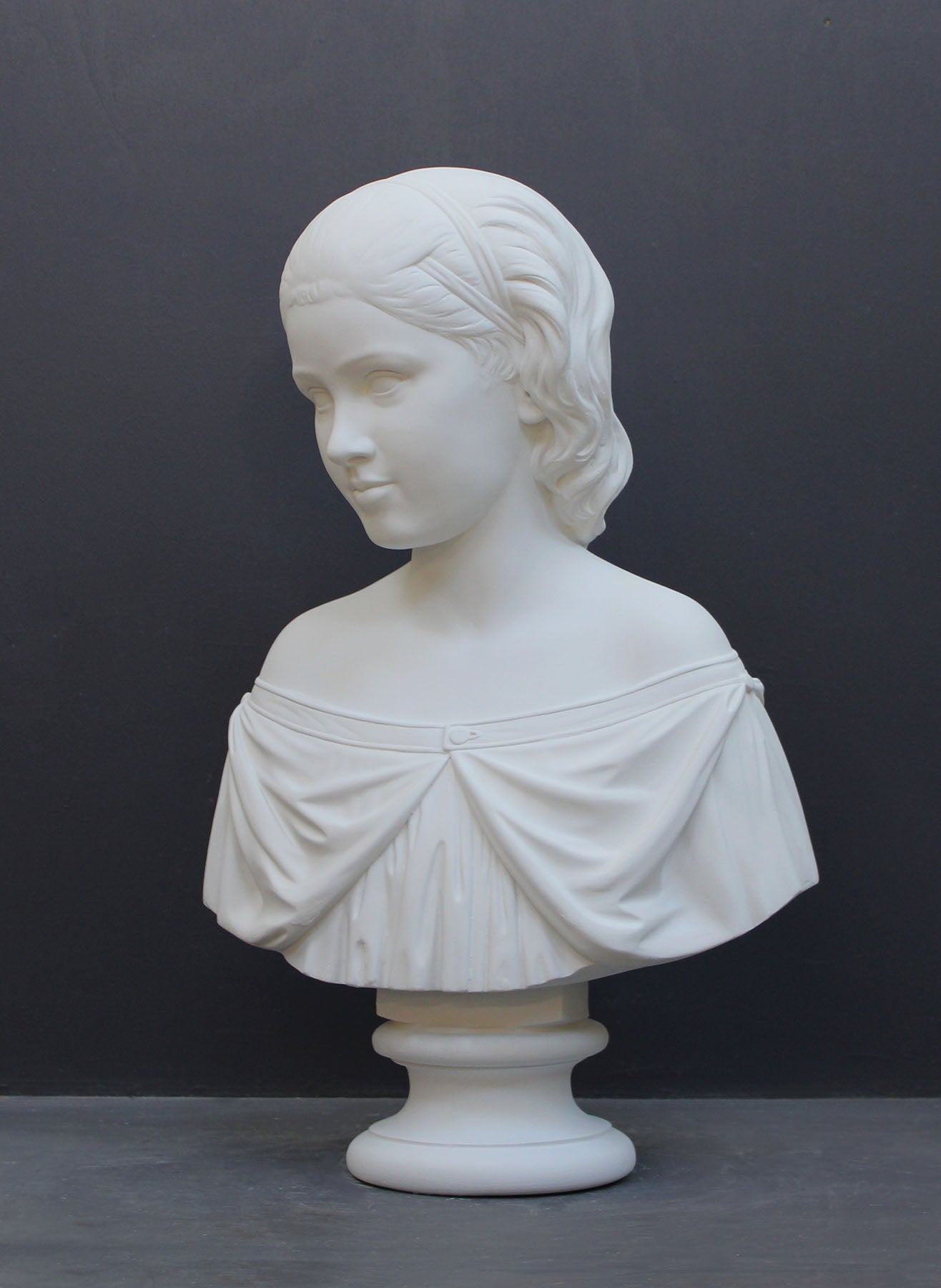 photo of white plaster cast bust sculpture of young woman with headband piece and in dress that falls off shoulders against a gray background