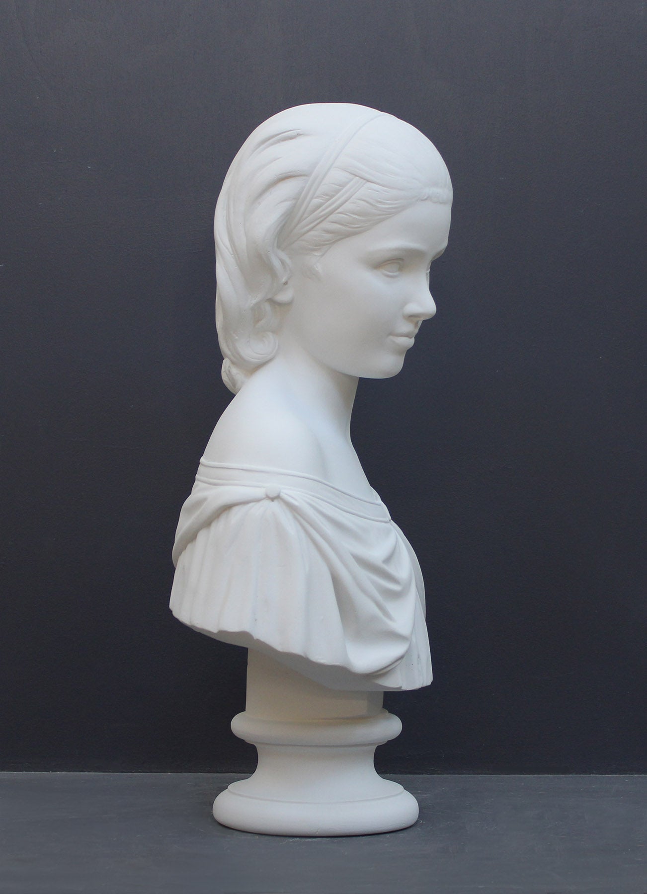 photo of white plaster cast bust sculpture of young woman with headband piece and in dress that falls off shoulders against a gray background