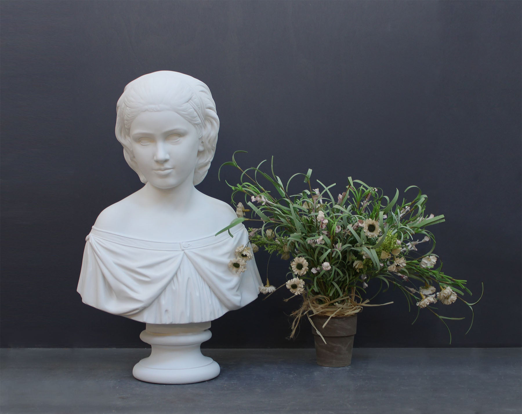 photo of white plaster cast bust sculpture of young woman with headband piece and in dress that falls off shoulders beside a white and pink flower arrangement against a gray background