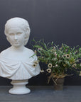 photo of white plaster cast bust sculpture of young woman with headband piece and in dress that falls off shoulders beside a white and pink flower arrangement against a gray background