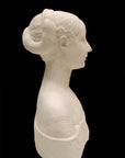 side view photo of white plaster cast sculpture of female bust cropped at shoulders with decorative base against black background