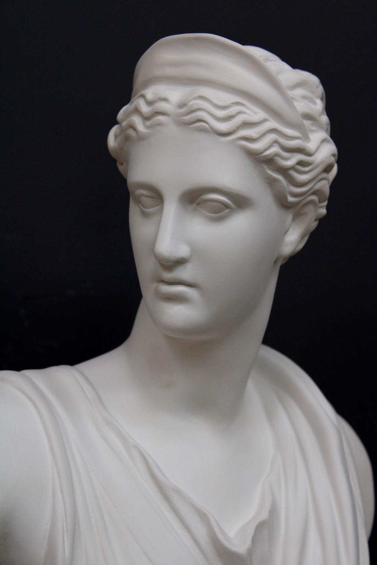 closeup photo of white plaster cast bust sculpture of woman, namely the goddess Diana, with drapery and crown and up-do against a black background