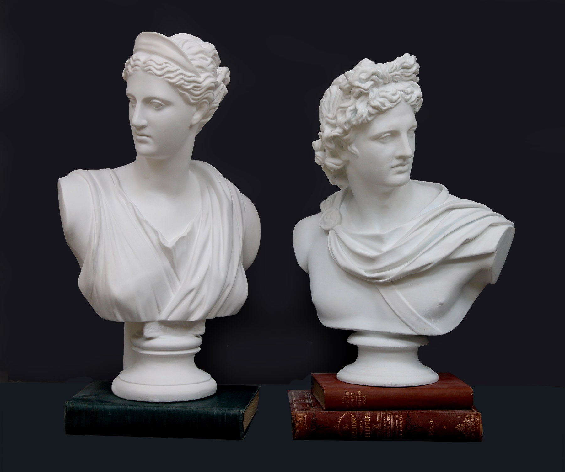 photo of two white plaster cast bust sculptures, one on right being a man, namely the god Apollo, with hair piled high in the front and a broach near his neck holding robes set atop two stacked red books, and other bust on the left being a woman, namely the goddess Diana, with drapery and crown and up-do set atop a green book, all against a dark gray background