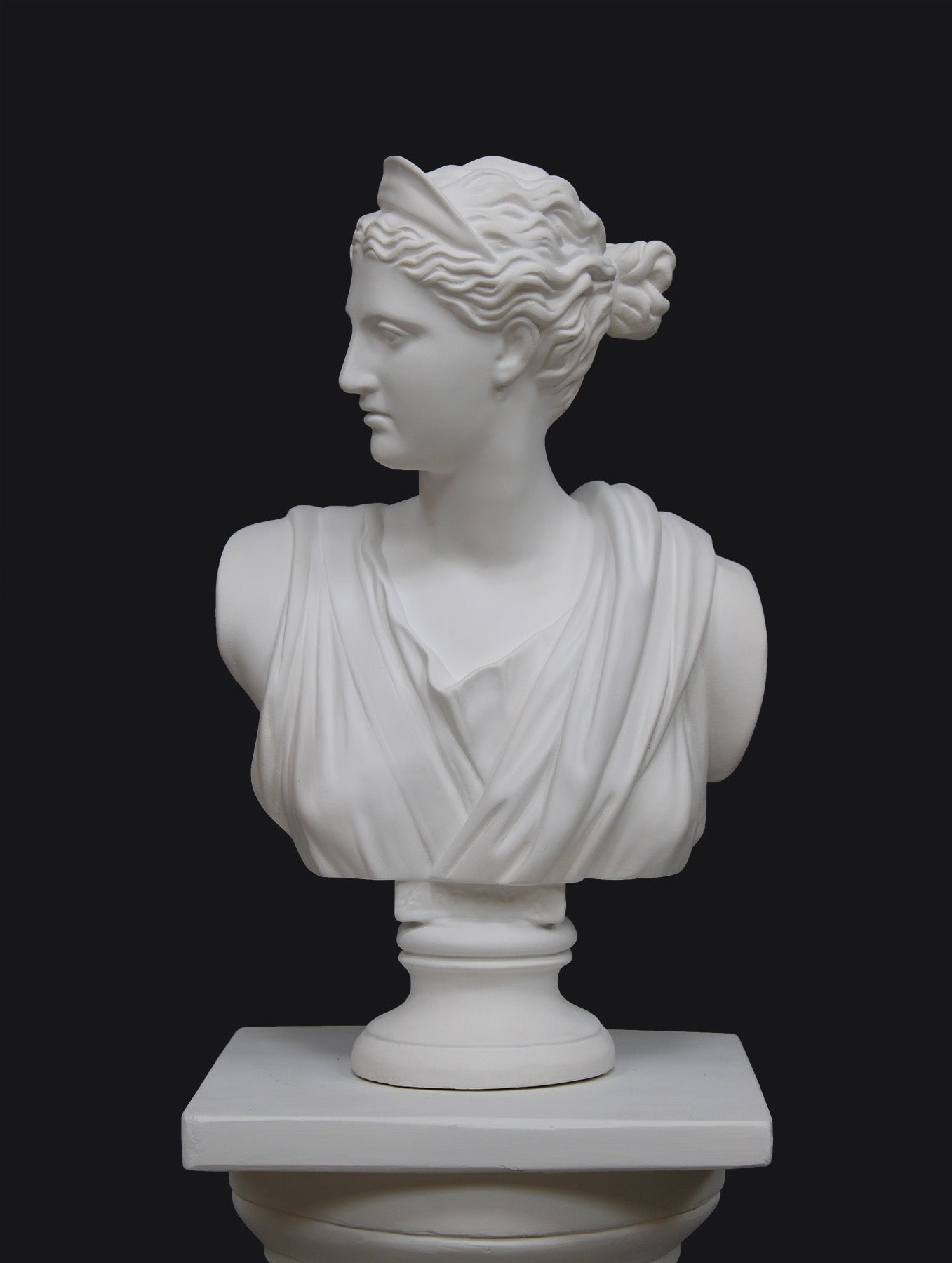 photo of white plaster cast bust sculpture of woman, namely the goddess Diana, with drapery and crown and up-do set atop a short white plaster cast pedestal in the Doric style with a fluted column against a gray background