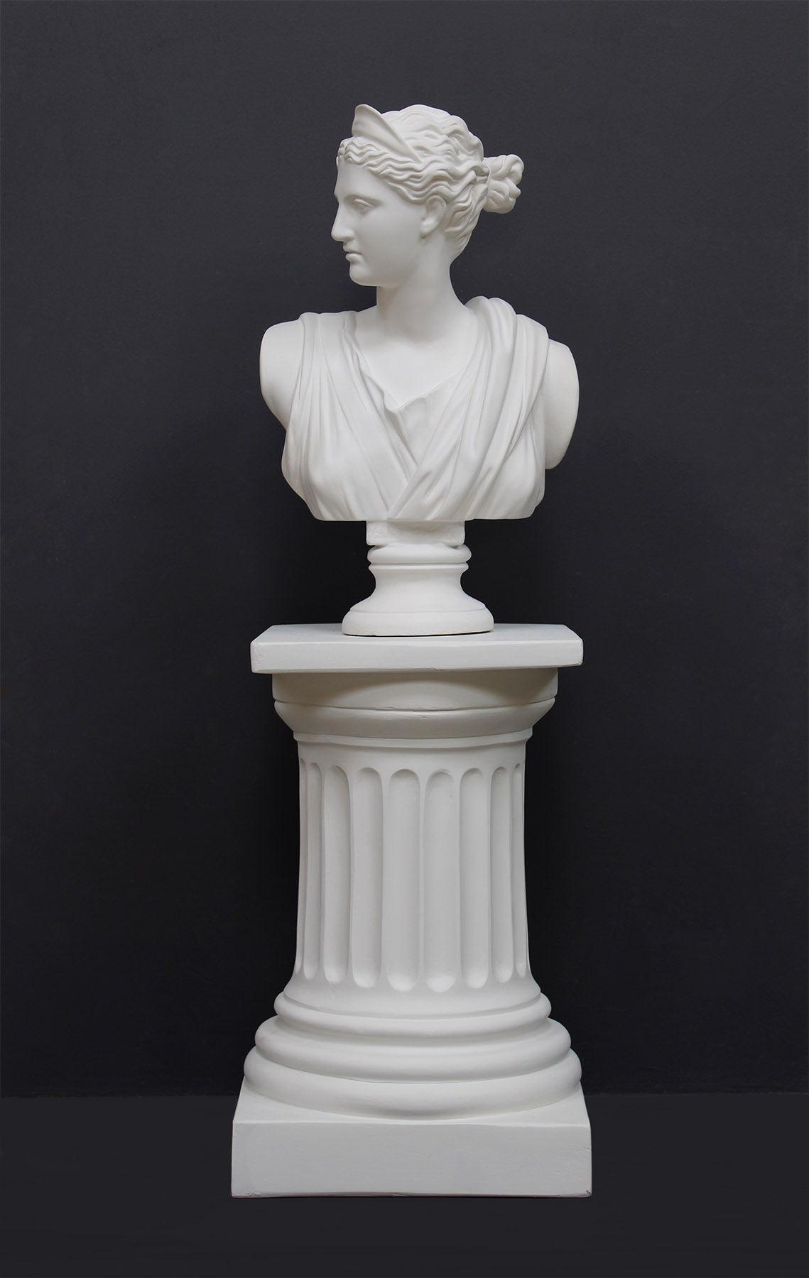 photo of white plaster cast bust sculpture of woman, namely the goddess Diana, with drapery and crown and up-do set atop a short white plaster cast pedestal in the Doric style with a fluted column against a gray background