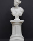 photo of white plaster cast bust sculpture of woman, namely the goddess Diana, with drapery and crown and up-do set atop a short white plaster cast pedestal in the Doric style with a fluted column against a gray background