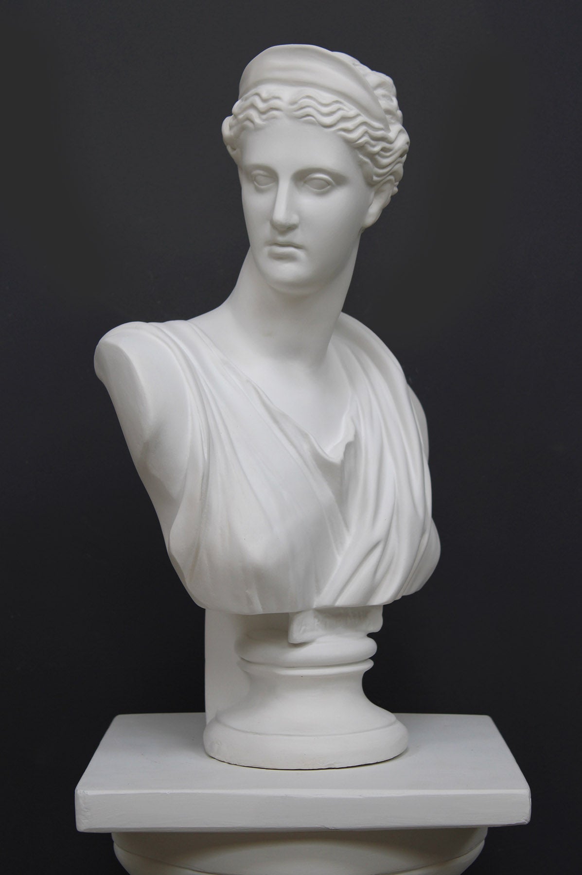 photo of white plaster cast bust sculpture of woman, namely the goddess Diana, with drapery and crown and up-do set atop a short white plaster cast pedestal in the Doric style with a fluted column against a gray background