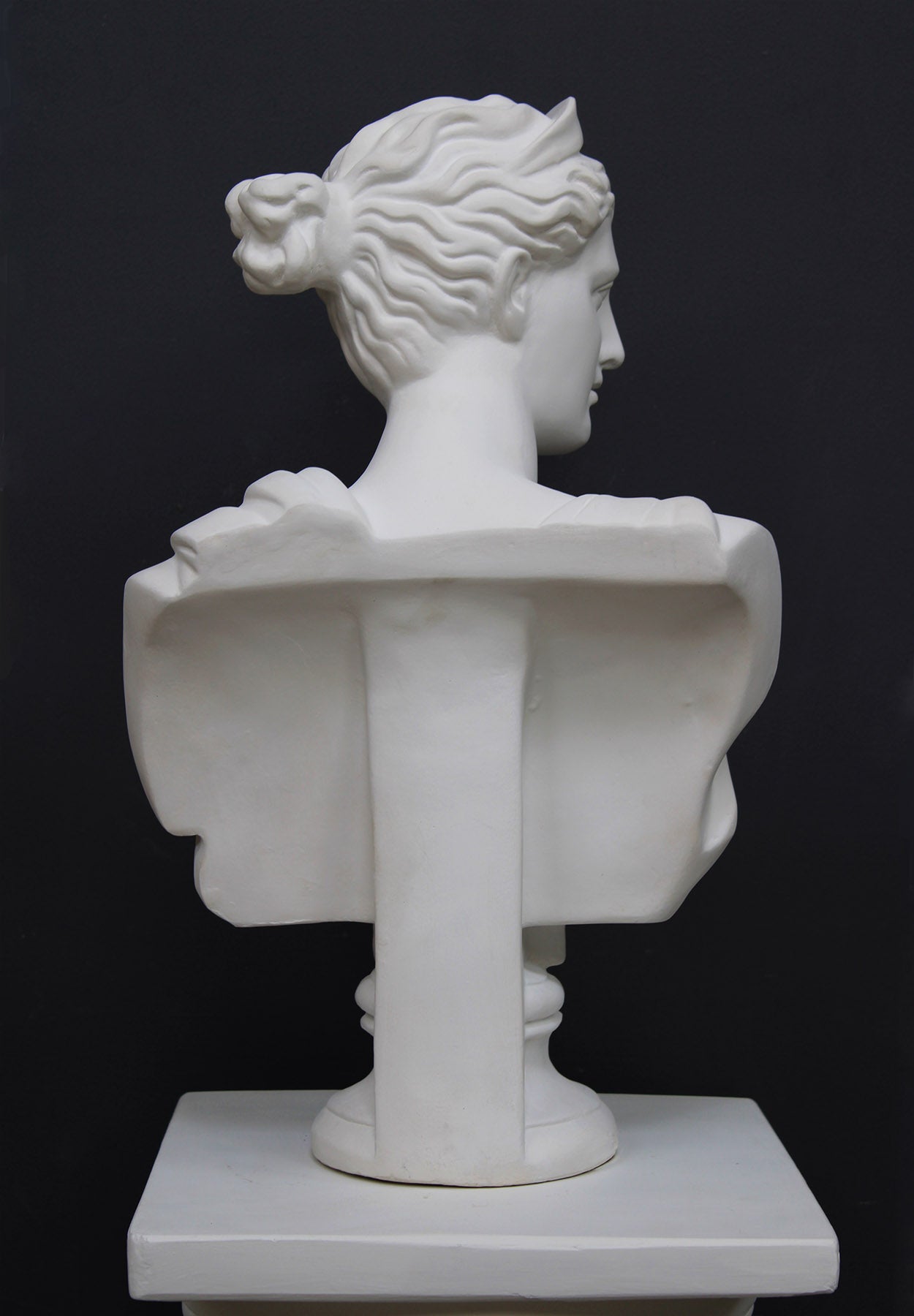 back view photo of white plaster cast bust sculpture of woman, namely the goddess Diana, with drapery and crown and up-do set atop a short white plaster cast pedestal in the Doric style with a fluted column against a gray background