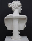 back view photo of white plaster cast bust sculpture of woman, namely the goddess Diana, with drapery and crown and up-do set atop a short white plaster cast pedestal in the Doric style with a fluted column against a gray background