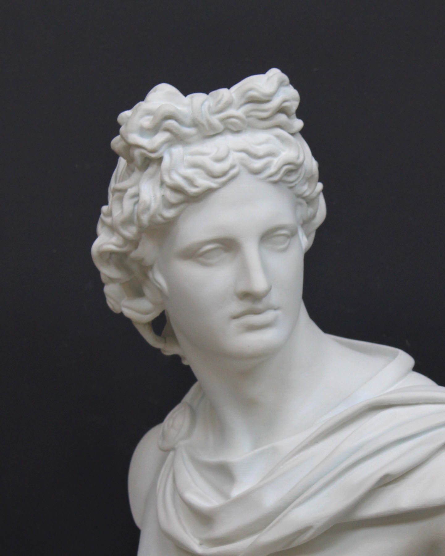 closeup photo of white plaster cast bust sculpture of man, namely the god Apollo, with hair piled high in the front and a broach near his neck holding robes against a gray background