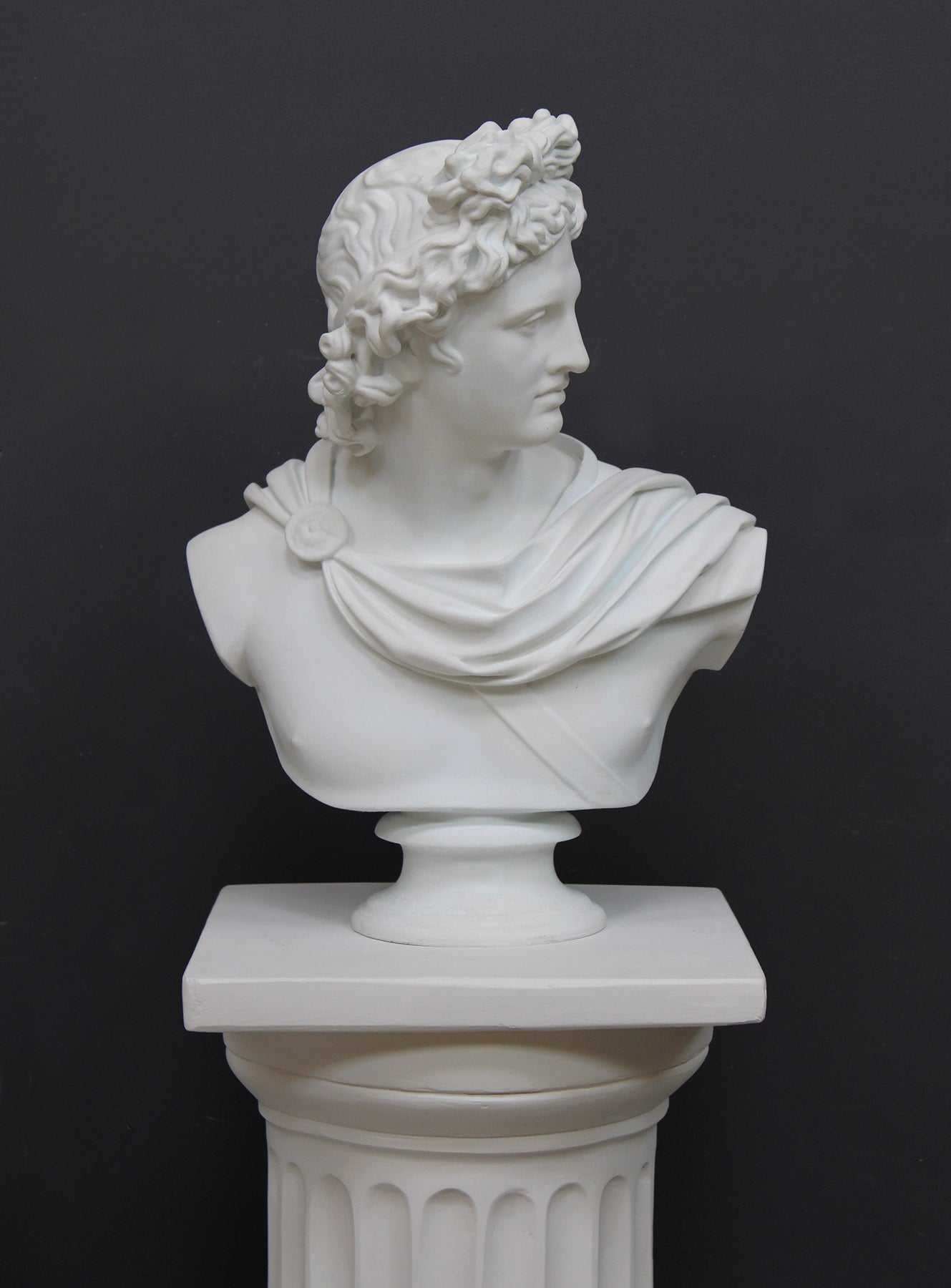 photo of white plaster cast bust sculpture of man, namely the god Apollo, with hair piled high in the front and a broach near his neck holding robes set atop a white pedestal against a gray background