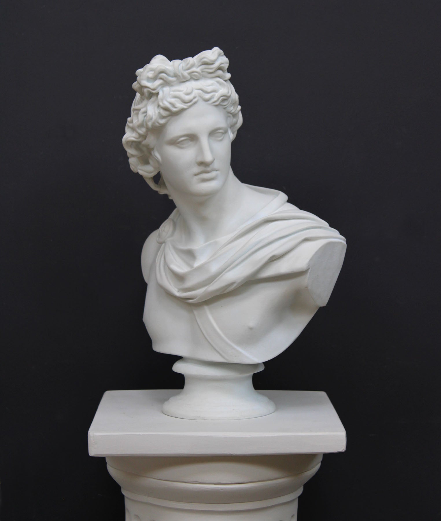 photo of white plaster cast bust sculpture of man, namely the god Apollo, with hair piled high in the front and a broach near his neck holding robes set atop a white pedestal against a gray background