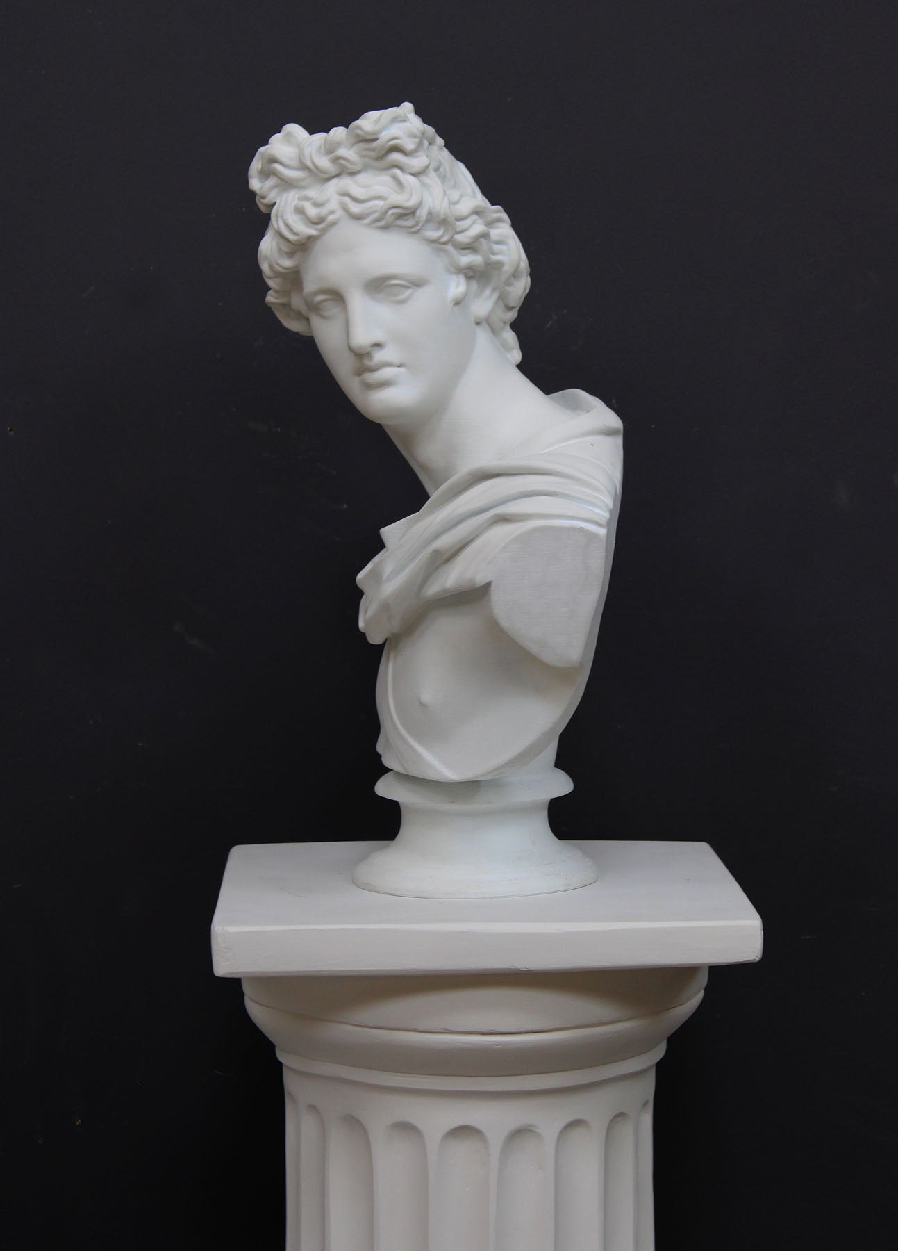 photo of white plaster cast bust sculpture of man, namely the god Apollo, with hair piled high in the front and a broach near his neck holding robes set atop a white pedestal against a gray background