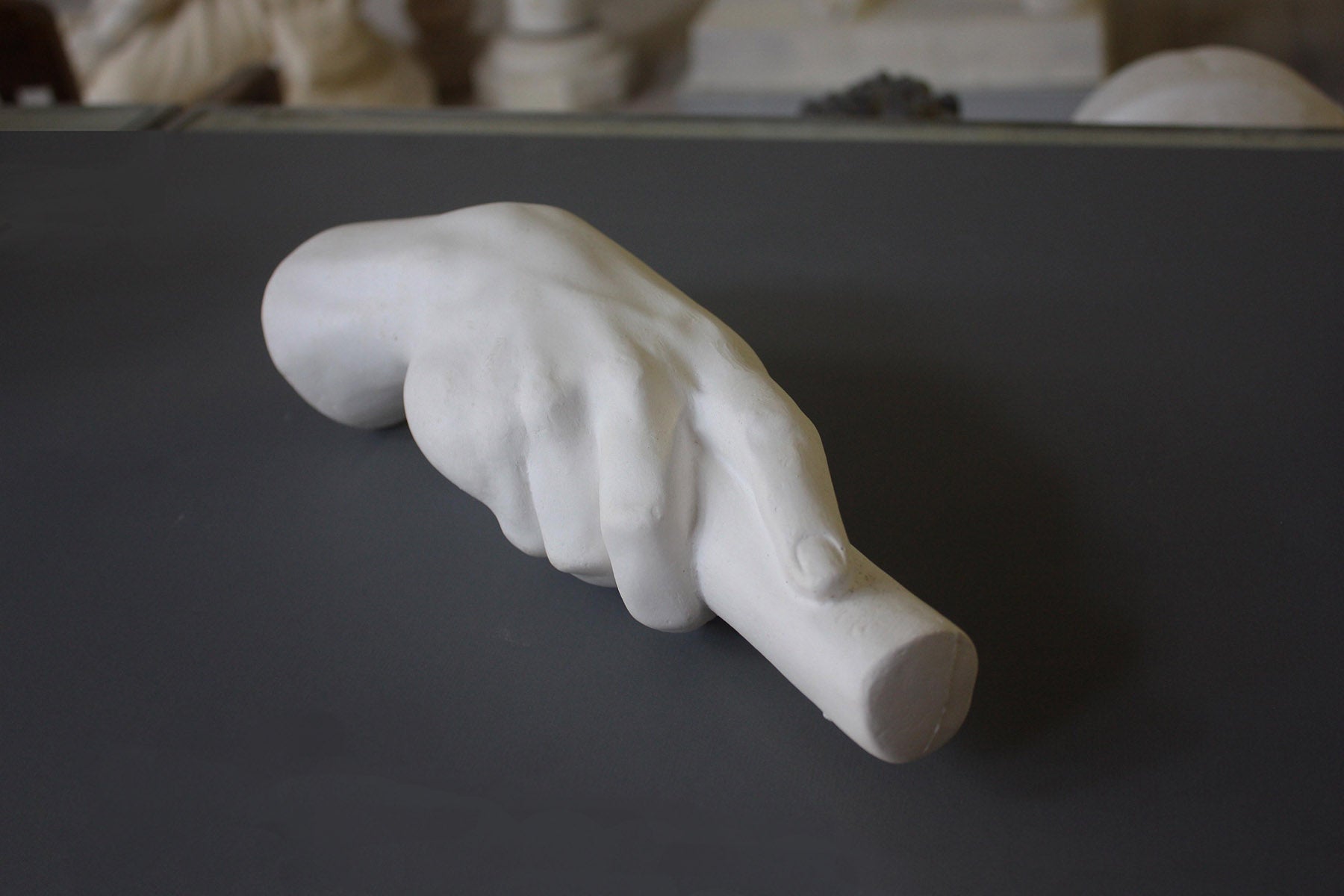 photo of white plaster cast of right hand grasping a staff or pole against gray surface with white plaster cast sculptures visible in background