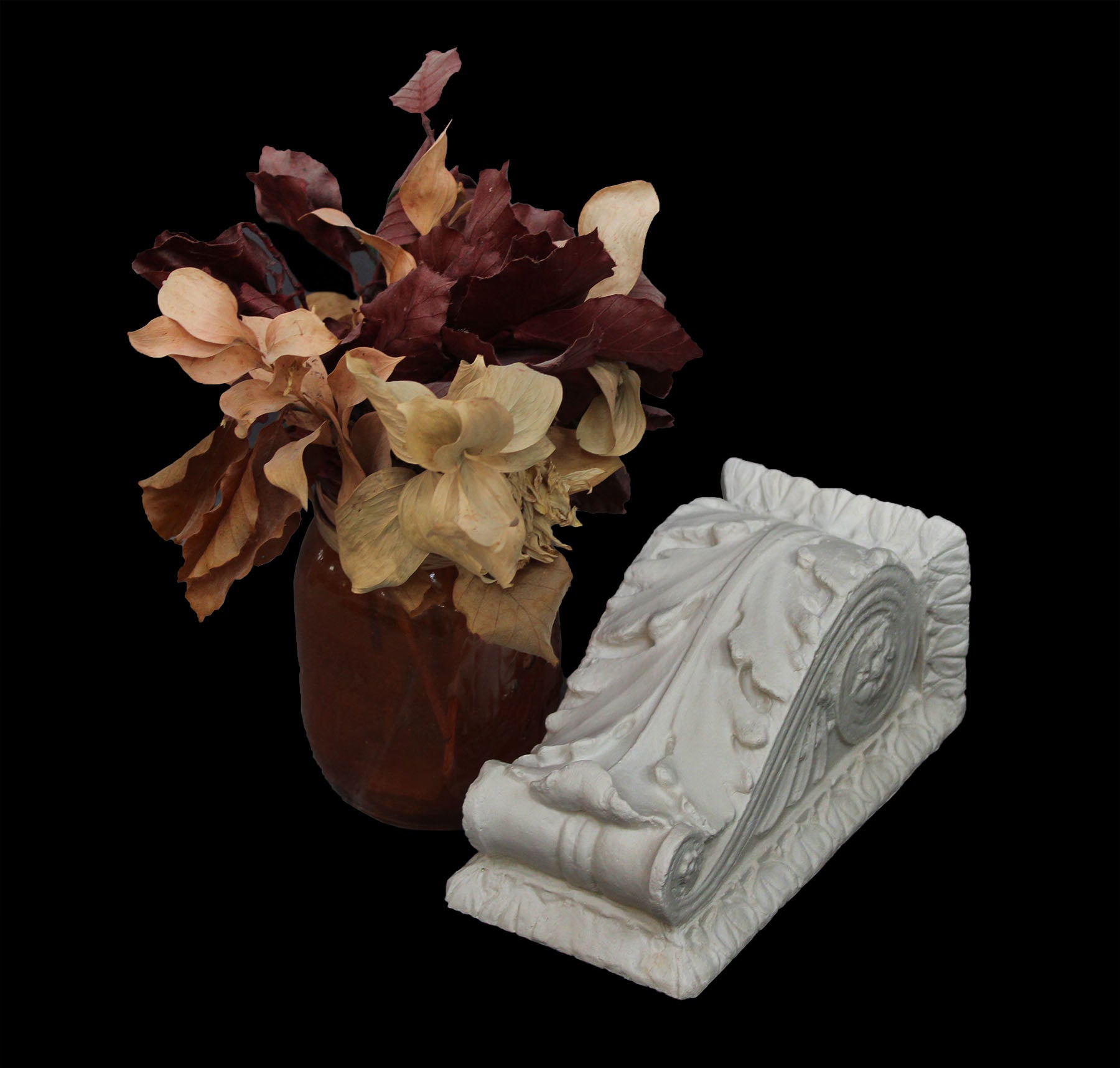 photo of white plaster cast sculpture of wall bracket with scrolls and acanthus leaves placed on a surface next to a red vase of red and off-white flowers against a black background