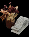 photo of white plaster cast sculpture of wall bracket with scrolls and acanthus leaves placed on a surface next to a red vase of red and off-white flowers against a black background