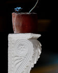 photo of white plaster cast sculpture of wall bracket with scrolls and acanthus leaves hung up with a red pot atop containing two blue flowers against a background of blacks, oranges, and dark blues