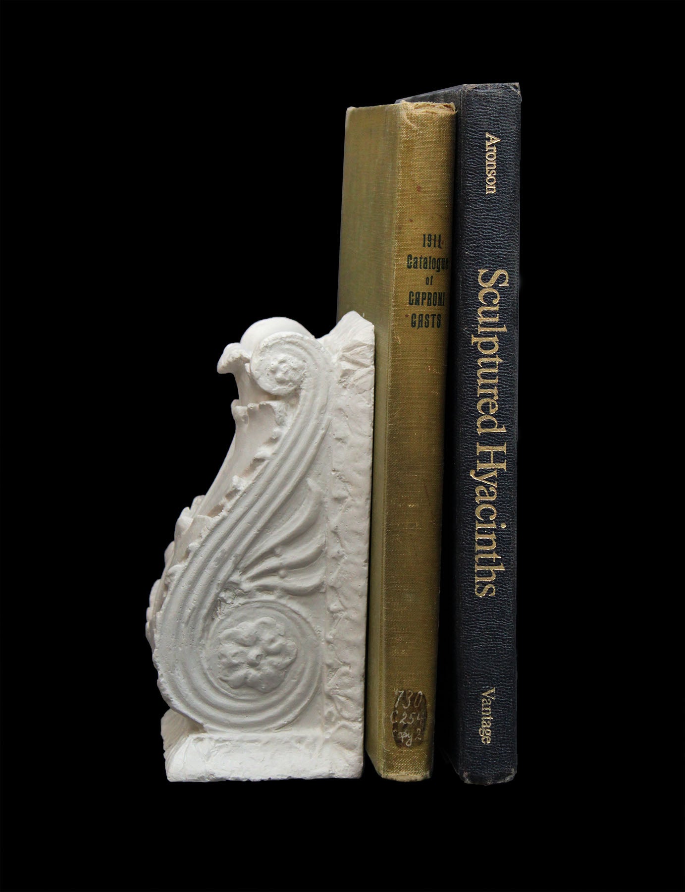 photo of white plaster cast sculpture of wall bracket with scrolls and acanthus leaves placed on a surface acting as a bookend with a green and black book beside it against a black background