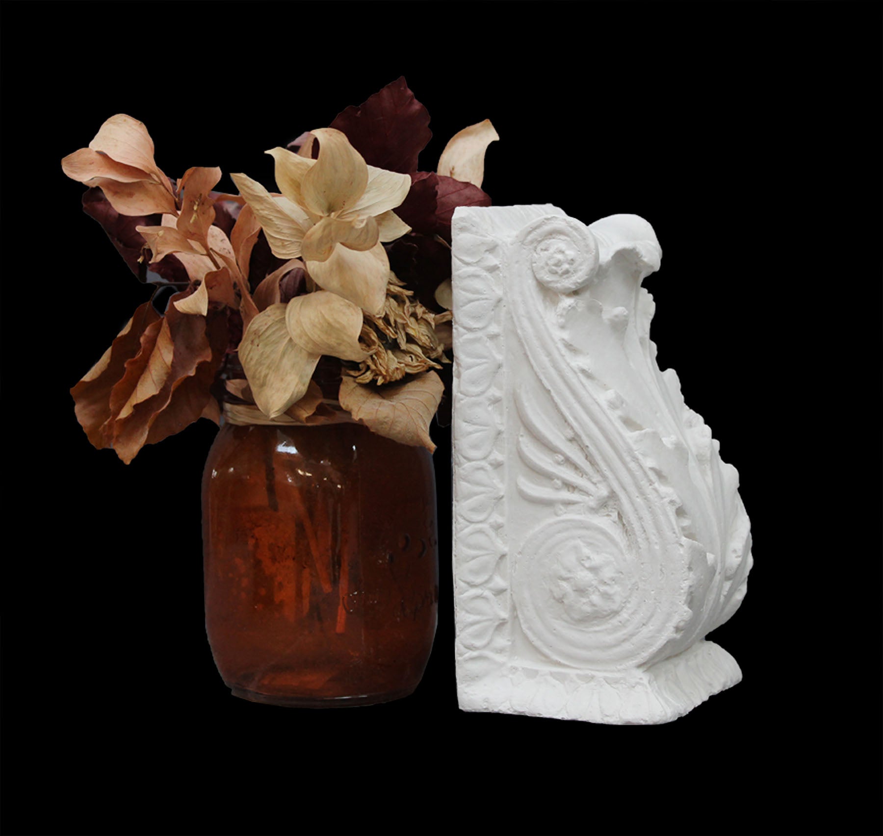 photo of white plaster cast sculpture of wall bracket with scrolls and acanthus leaves placed on a surface next to a red vase of red and off-white flowers against a black background